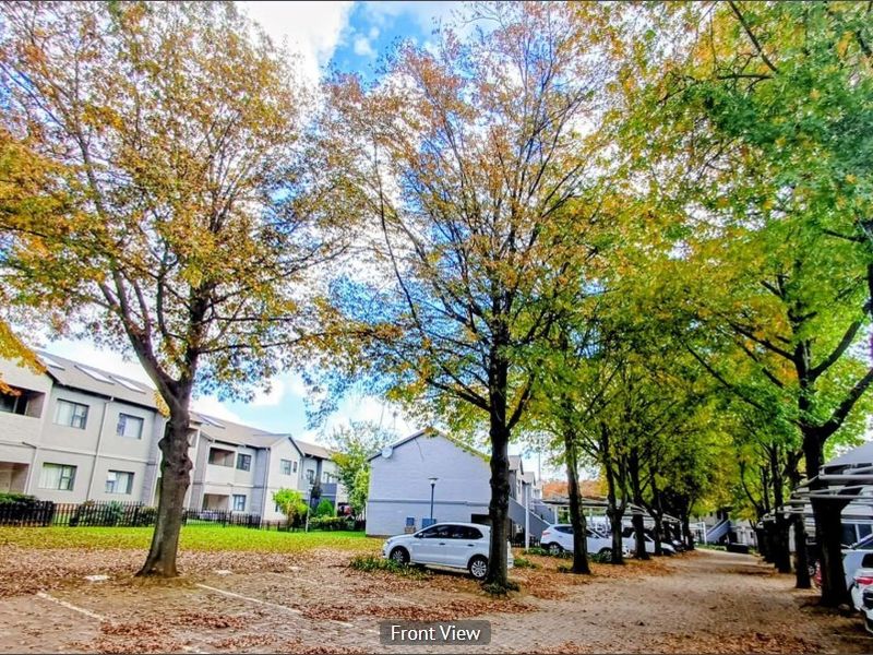 Exterior view of the apartment complex featuring a paved parking area lined with mature trees and building facades.