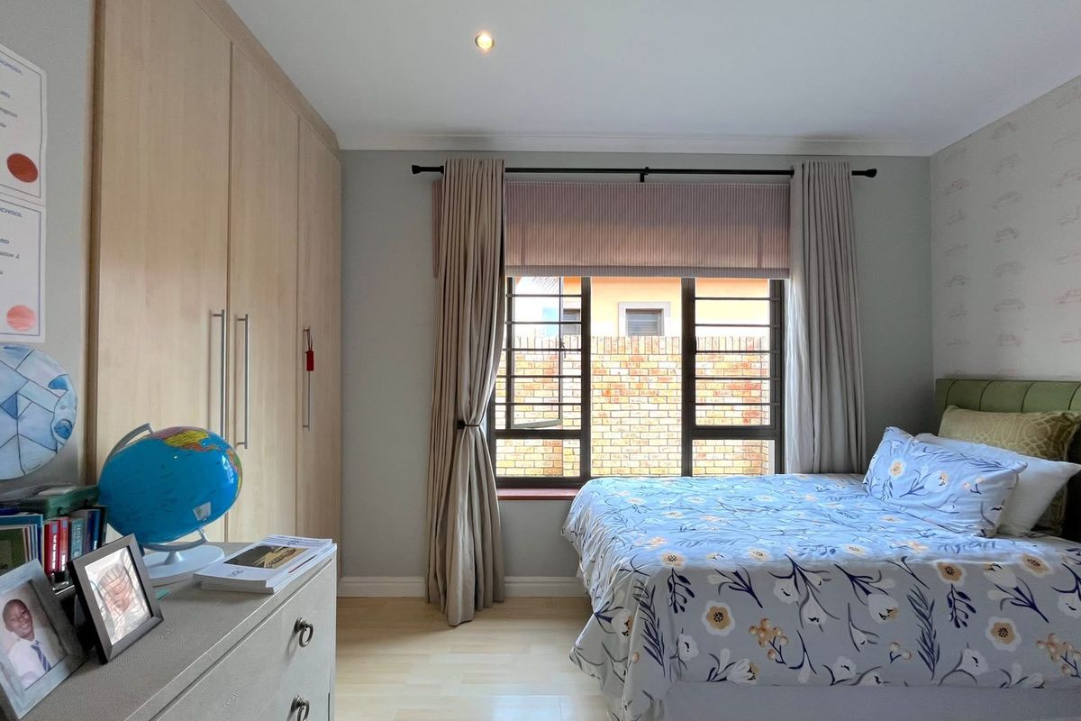 A well-maintained bedroom featuring light wood built-in cupboards, laminate flooring, and a window fitted with security bars and neutral curtains.
