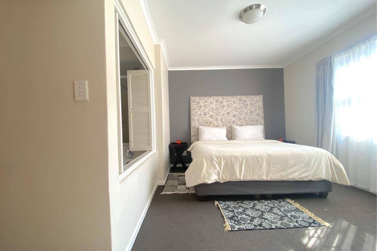 A well-maintained bedroom featuring carpeted flooring, a neutral color palette with a feature wall, and an internal window with shutters.