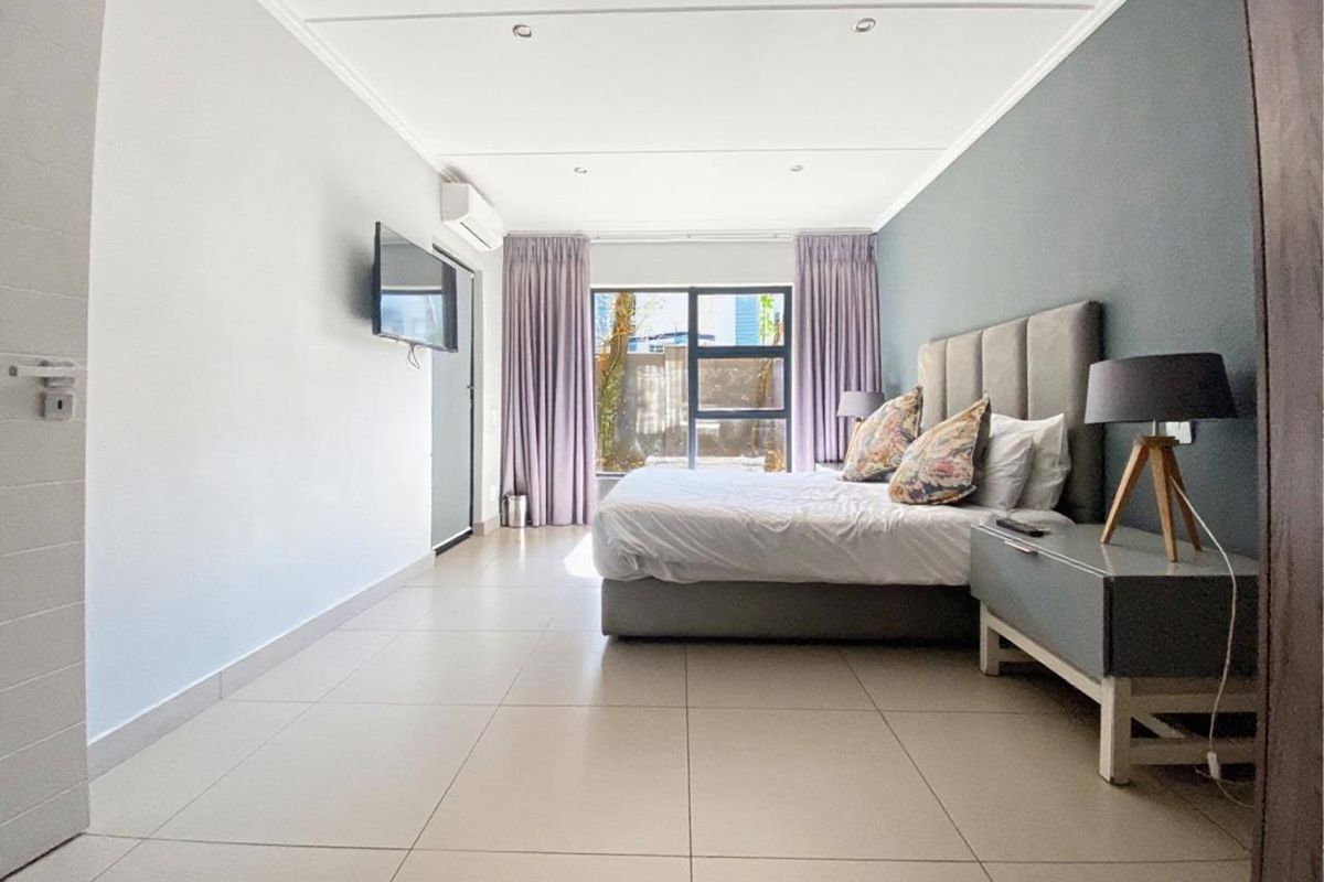 A spacious, modern bedroom featuring neutral tiled flooring, recessed lighting, a wall-mounted air conditioning unit, and large windows with natural light.