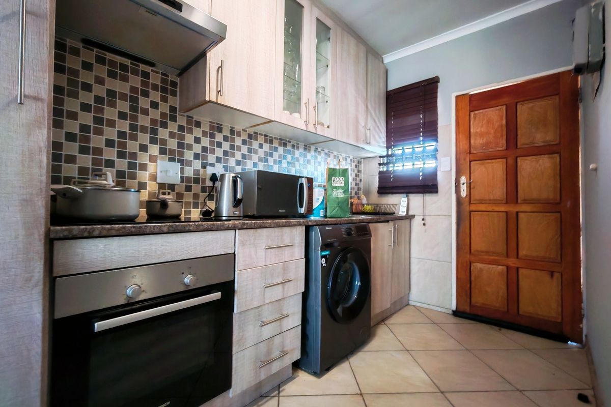 Compact kitchen featuring light wood-finish cabinetry, mosaic tile backsplash, built-in oven with extractor, and integrated laundry appliance, finished with ceramic floor tiles.