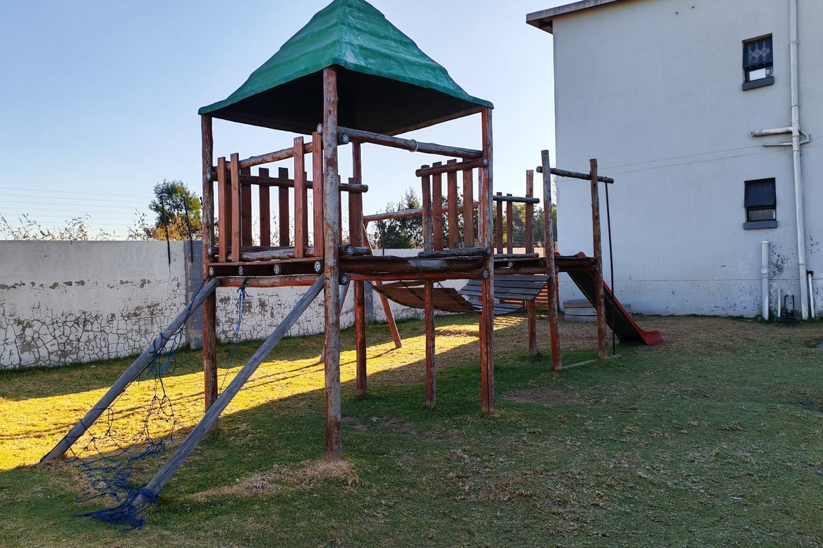Rear exterior area featuring a communal playground structure set on a grass lawn with a boundary wall and apartment building facade.