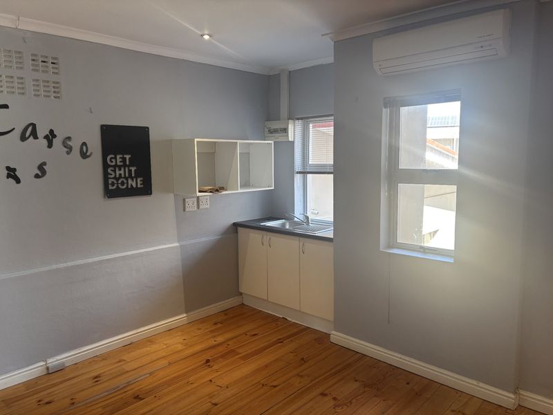 A compact kitchenette featuring white cabinetry, a stainless steel sink, timber flooring, and a wall-mounted air conditioning unit in a neutral-toned office space.