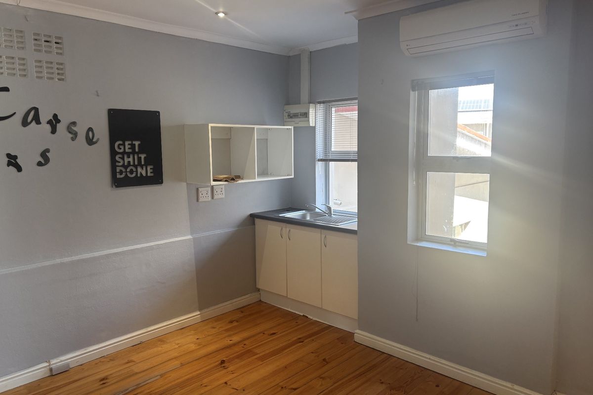 A compact kitchenette featuring white cabinetry, a stainless steel sink, timber flooring, and a wall-mounted air conditioning unit in a neutral-toned office space.