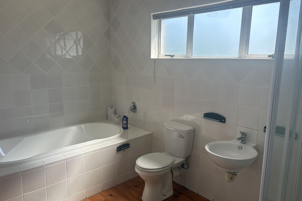 A functional bathroom featuring a built-in bathtub, wall-mounted toilet, and basin, finished with white wall tiles and timber flooring.