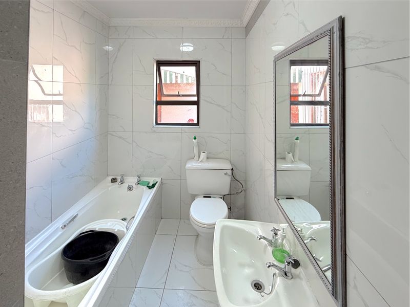A clean, modern bathroom featuring marble-effect wall tiles, a white bathtub, a pedestal basin, and a wall-mounted mirror.