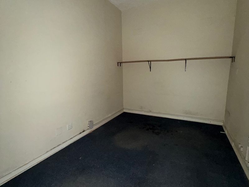 An empty, dated office space featuring dark carpeting, wall-mounted shelving, and surface-mounted electrical infrastructure, requiring cosmetic updates and refurbishment.