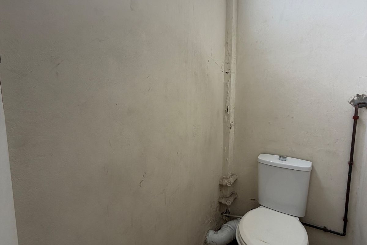 A small, functional bathroom space featuring a toilet, tiled flooring, and a high-level window, currently requiring cosmetic updates and wall repairs.