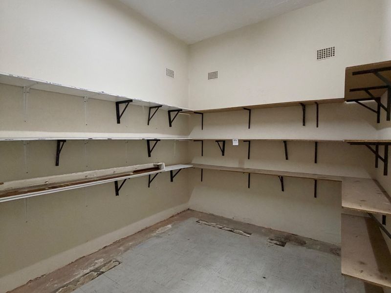 A vacant utility or storage room featuring extensive built-in wall shelving, fluorescent lighting, and worn flooring, suitable for various industrial storage applications.