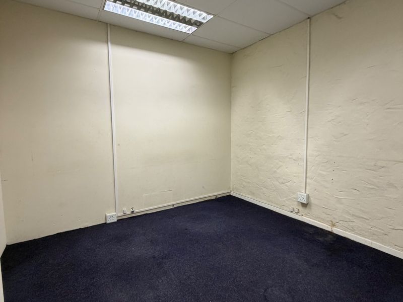 An empty office space featuring a suspended ceiling with recessed lighting, surface-mounted electrical conduits, and dark carpet flooring.