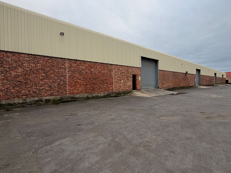 Rear exterior view of an industrial warehouse featuring brick and metal cladding, multiple roller shutter doors, and a large asphalt yard area.