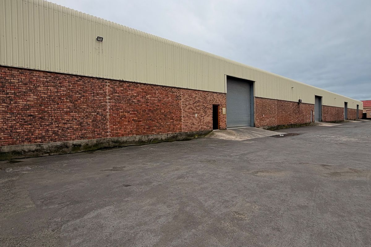Rear exterior view of an industrial warehouse featuring brick and metal cladding, multiple roller shutter doors, and a large asphalt yard area.