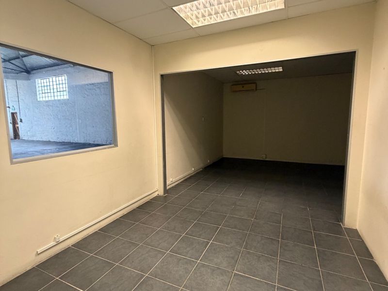 An empty office space featuring tiled flooring, a suspended ceiling with recessed lighting, and an internal window overlooking the warehouse area.