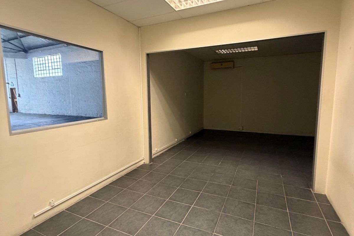 An empty office space featuring tiled flooring, a suspended ceiling with recessed lighting, and an internal window overlooking the warehouse area.