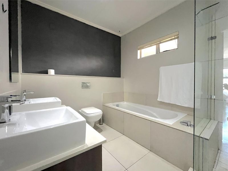 Modern bathroom featuring a double vanity, built-in bathtub, wall-mounted toilet, and a glass-enclosed shower with neutral tiled finishes.