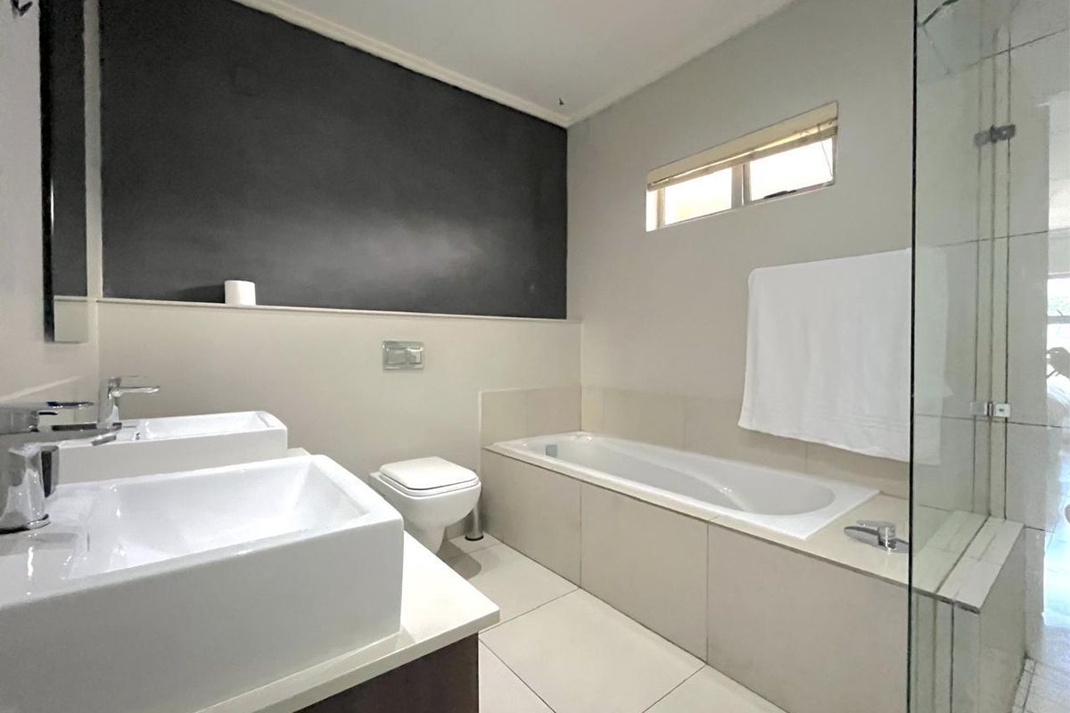 Modern bathroom featuring a double vanity, built-in bathtub, wall-mounted toilet, and a glass-enclosed shower with neutral tiled finishes.