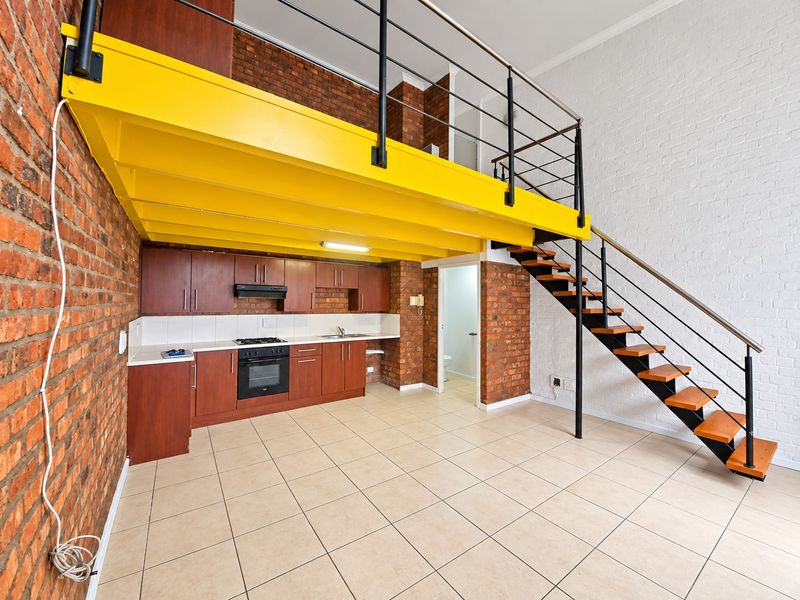 Open-plan living area featuring a kitchen with built-in cabinetry, exposed brick walls, and a mezzanine level accessed by a metal staircase.