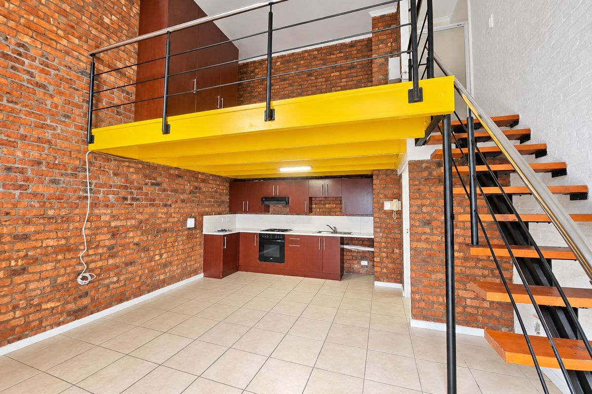 Open-plan kitchen and living area featuring exposed brick walls, a mezzanine level with a steel staircase, and functional built-in cabinetry.