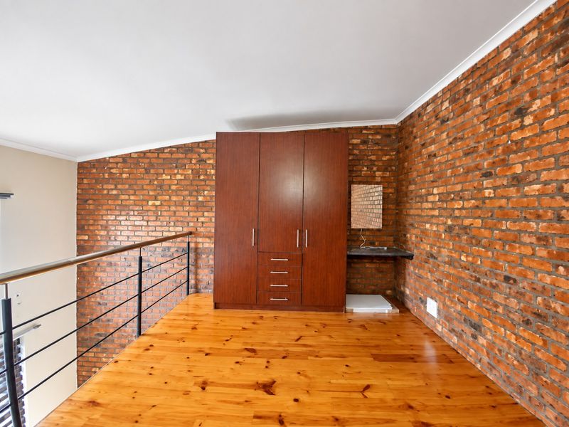 A loft-style bedroom featuring warm timber flooring, exposed brick walls, and a built-in wardrobe with an integrated floating desk.