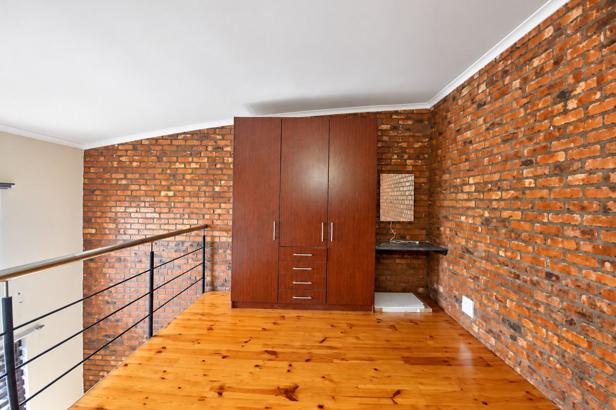 A loft-style bedroom featuring warm timber flooring, exposed brick walls, and a built-in wardrobe with an integrated floating desk.