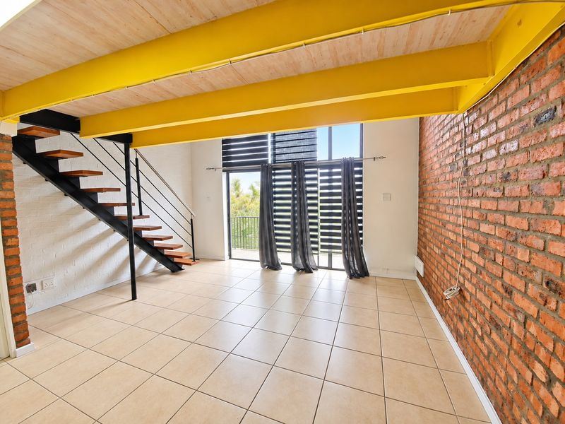 Spacious open-plan living area featuring exposed brick walls, industrial-style yellow steel beams, a modern staircase, and large sliding doors leading to a balcony.