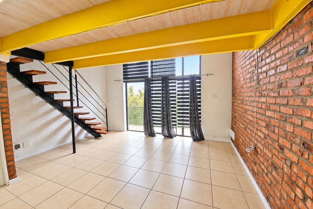 Spacious open-plan living area featuring exposed brick walls, industrial-style yellow steel beams, a modern staircase, and large sliding doors leading to a balcony.