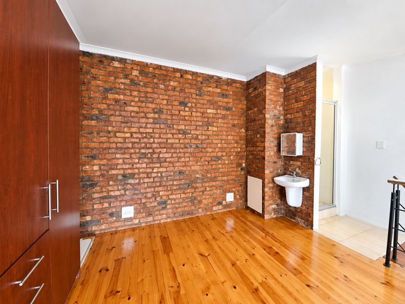 A bright bedroom featuring an exposed brick feature wall, polished hardwood flooring, a built-in wardrobe, and an en-suite shower area with a wall-mounted basin.