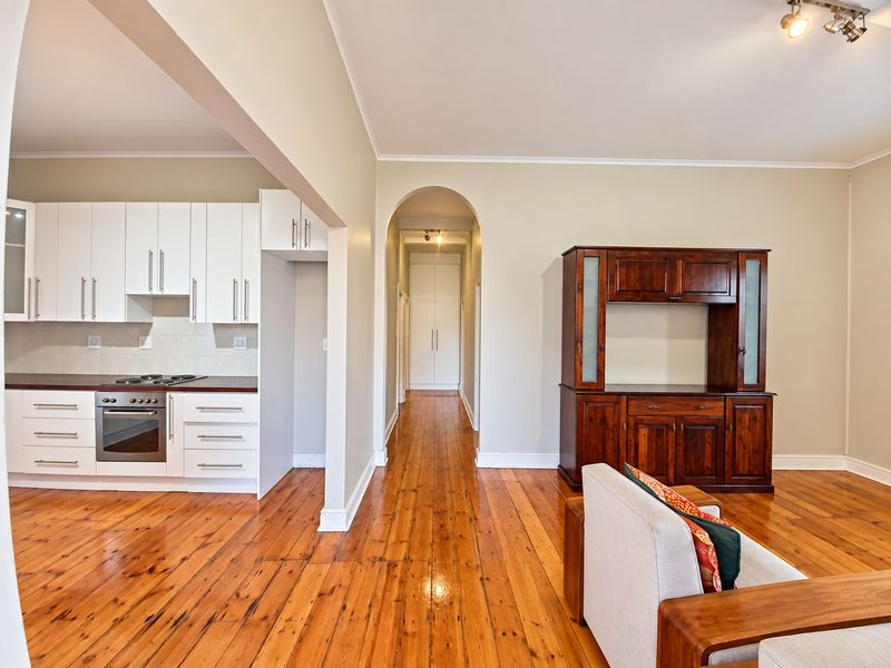 Open plan living area featuring polished hardwood floors, a modern white kitchen with built-in appliances, and an arched hallway leading to storage cupboards.