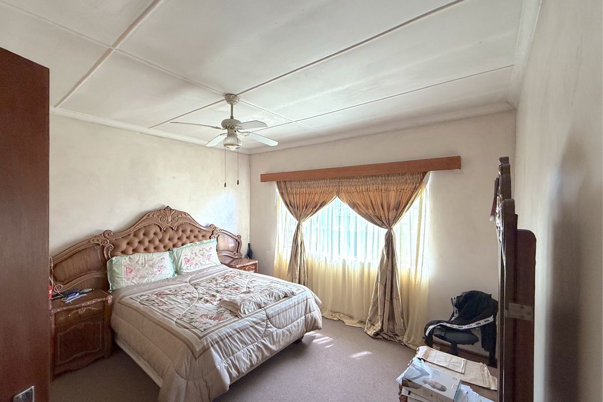 A bedroom featuring carpeted flooring, a ceiling fan, and a window with a wooden pelmet. The room shows signs of wear on the walls and ceiling.