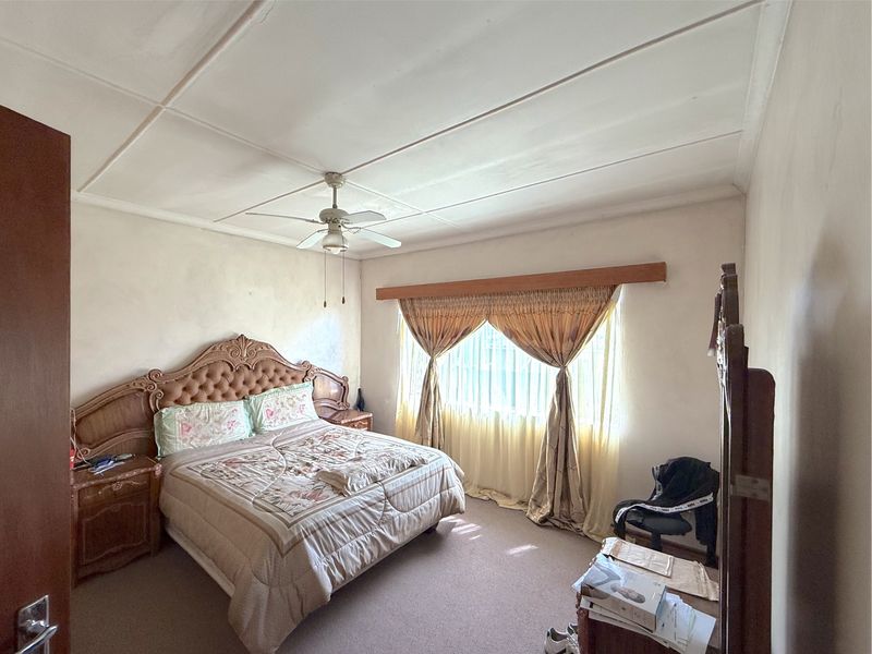 A bedroom featuring carpeted flooring, a ceiling fan, and a window with a wooden pelmet. The room shows signs of wear on the walls and ceiling.