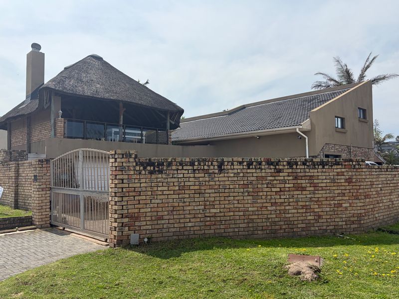 Front exterior view of a multi-level house featuring a combination of thatch and tiled roofing, enclosed by a brick boundary wall and metal gate.