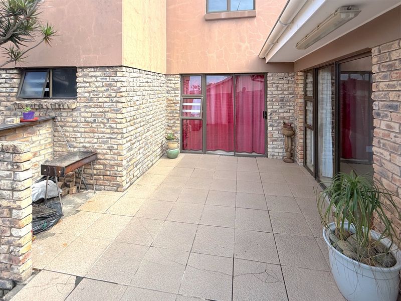 A paved outdoor patio area featuring a built-in brick braai, sliding door access, and exposed brick walls, requiring some maintenance to the flooring and plaster.