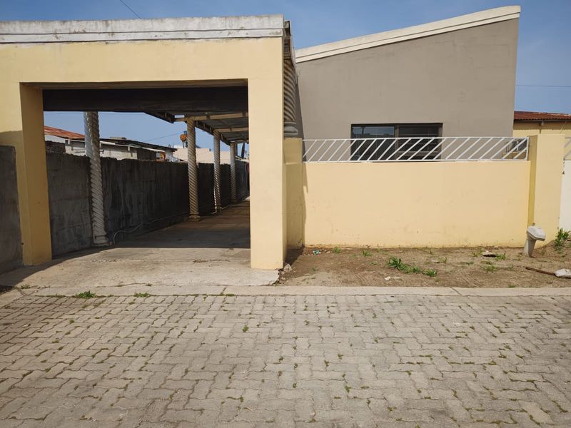Front exterior view featuring a covered carport, paved driveway, and boundary wall with security fencing, showing signs of weathering and maintenance requirements.