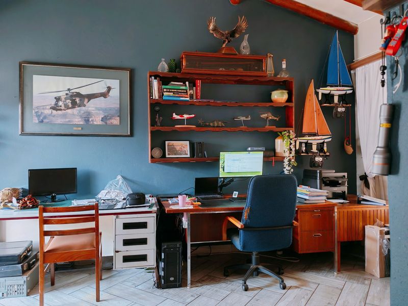 A home office space featuring dark blue walls, built-in shelving, and tiled flooring, providing a functional workspace with exposed timber ceiling details.