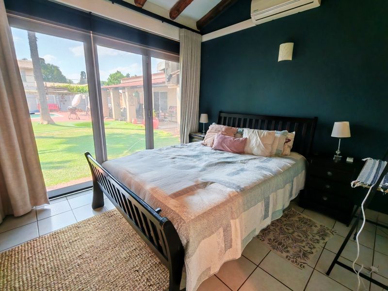 A well-lit bedroom featuring exposed timber ceiling beams, tiled flooring, and large sliding glass doors providing direct access to the garden and pool area.