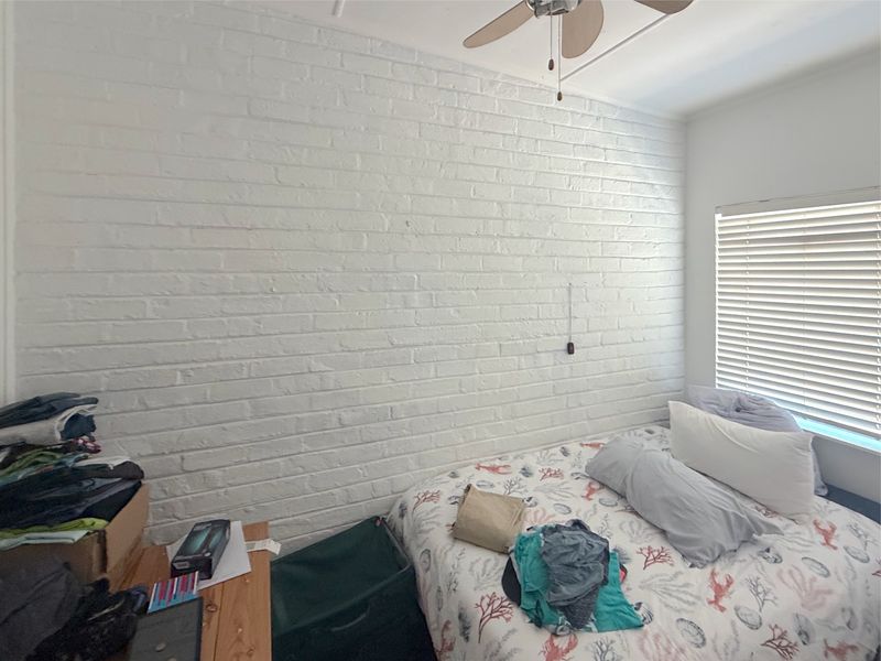 A bedroom featuring a white painted brick accent wall, a ceiling fan, and venetian blinds over the window.