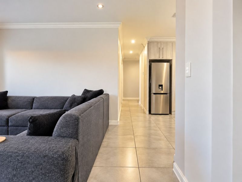 An open-plan living area featuring neutral tiled flooring, recessed downlighting, and a seamless transition into a hallway with built-in cabinetry.