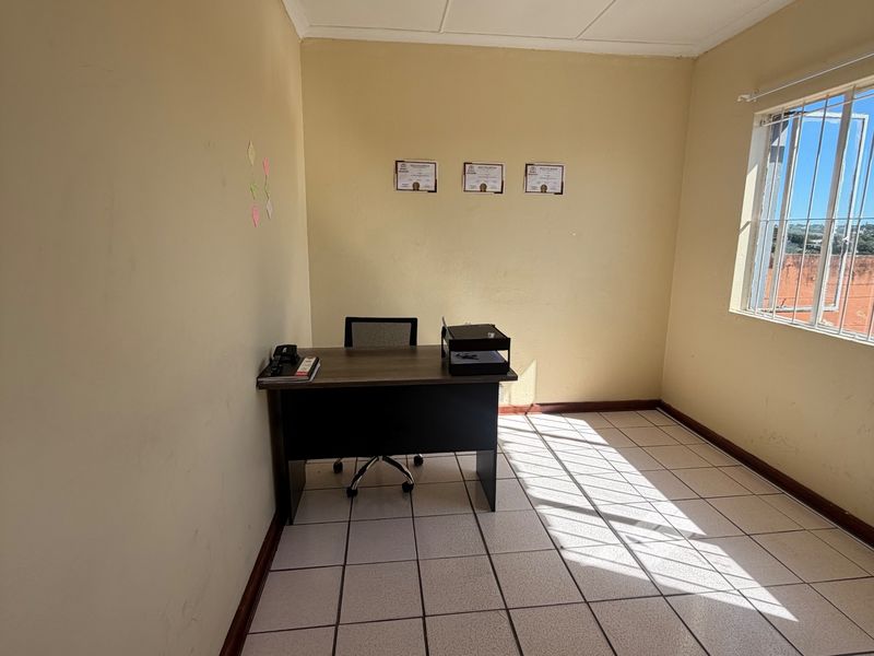 A functional office space featuring ceramic floor tiles, neutral wall finishes, and a window equipped with security bars, providing natural light.