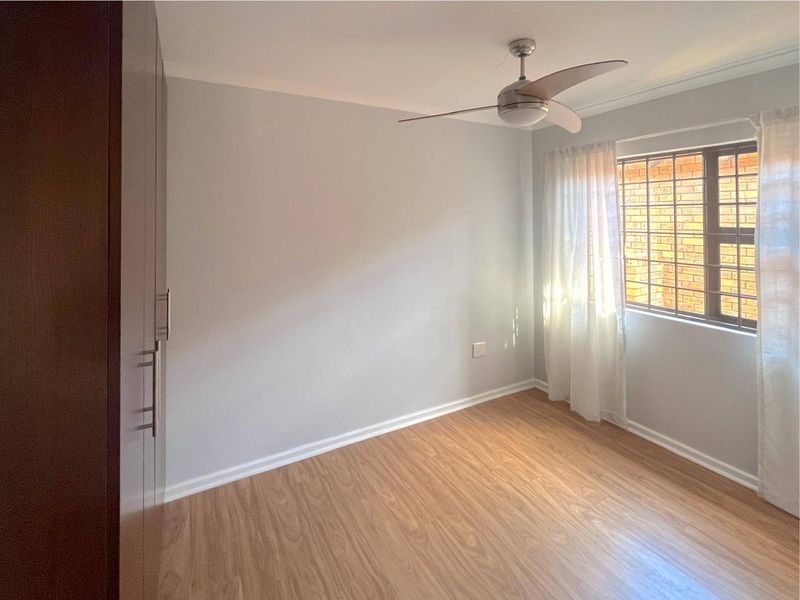 A bright, empty bedroom featuring laminate flooring, built-in cupboards, and a ceiling fan, with security bars installed on the window.