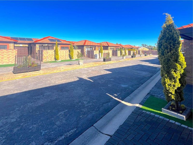 A view of a residential townhouse complex street featuring uniform brick facades, tiled roofs, solar panels, and paved driveways.