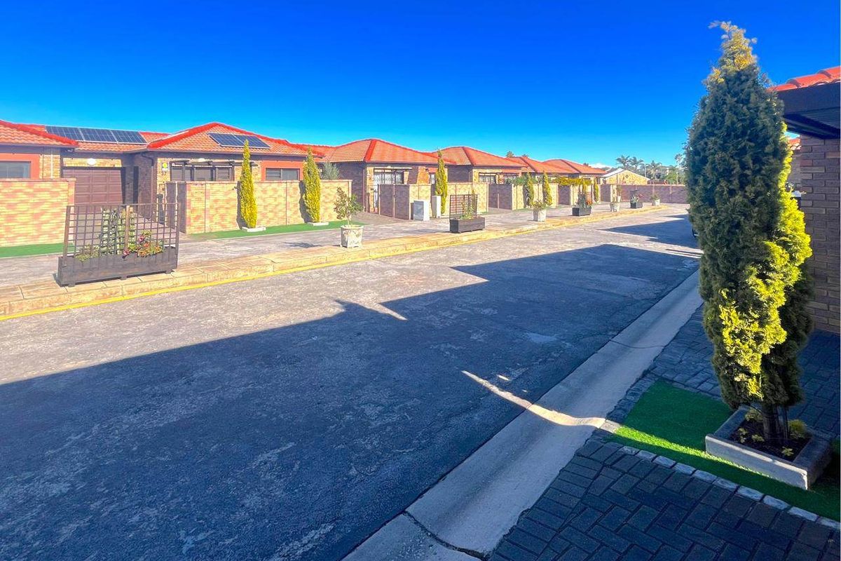 A view of a residential townhouse complex street featuring uniform brick facades, tiled roofs, solar panels, and paved driveways.