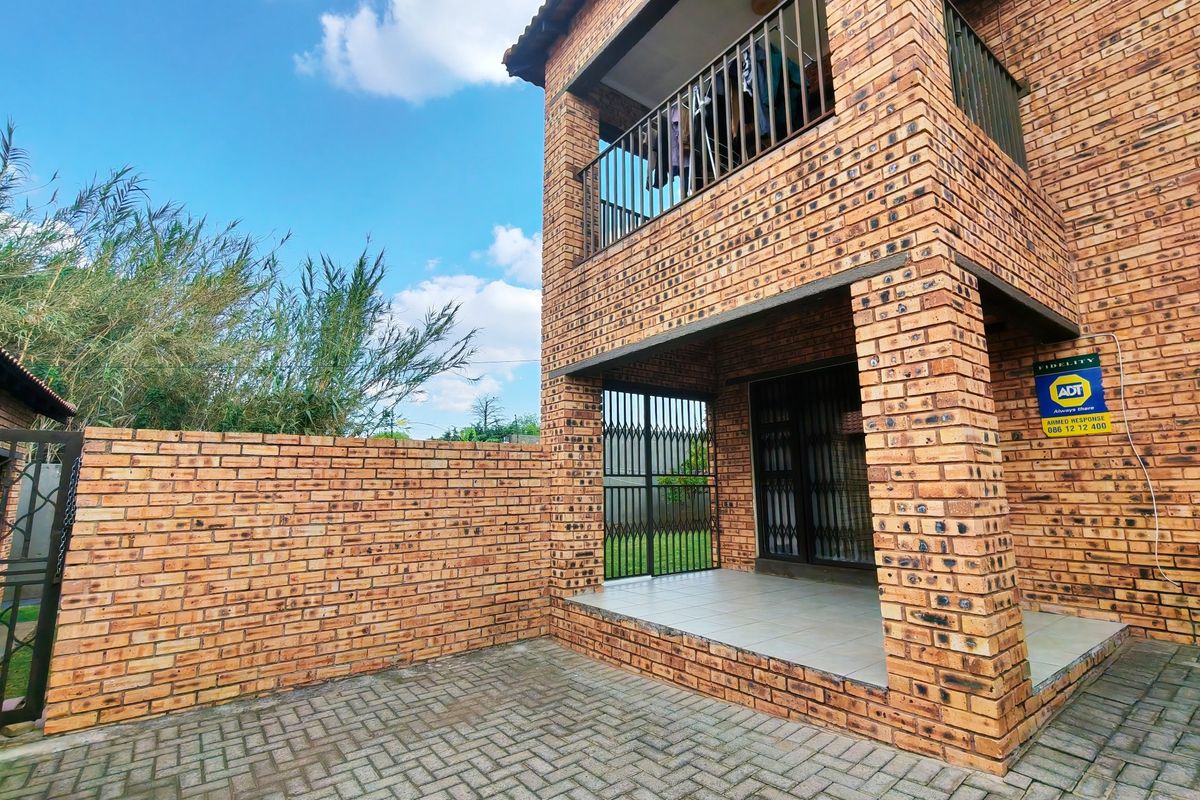 Rear view of a double-story face brick apartment featuring a ground-floor tiled patio, upper-level balcony, and a private paved courtyard with boundary walling.