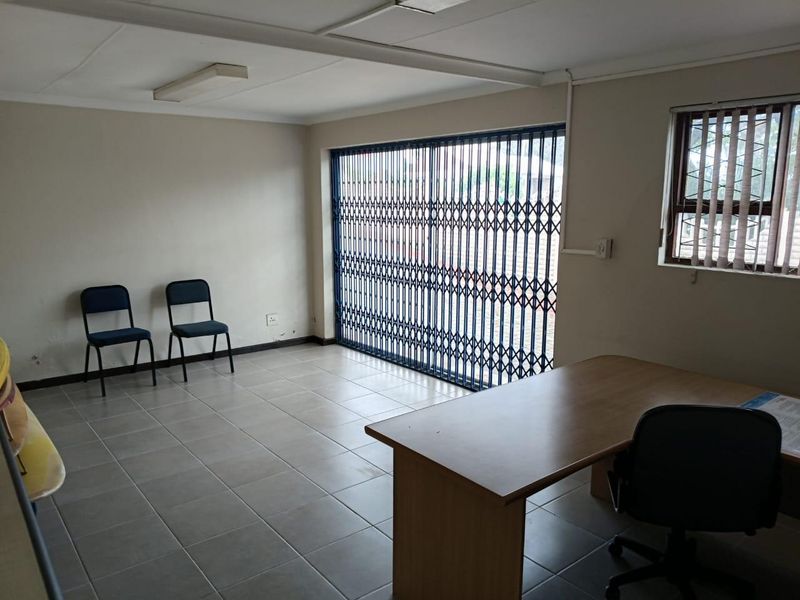 A functional commercial office space featuring tiled flooring, a secure sliding gate, and natural light through a window with security bars.