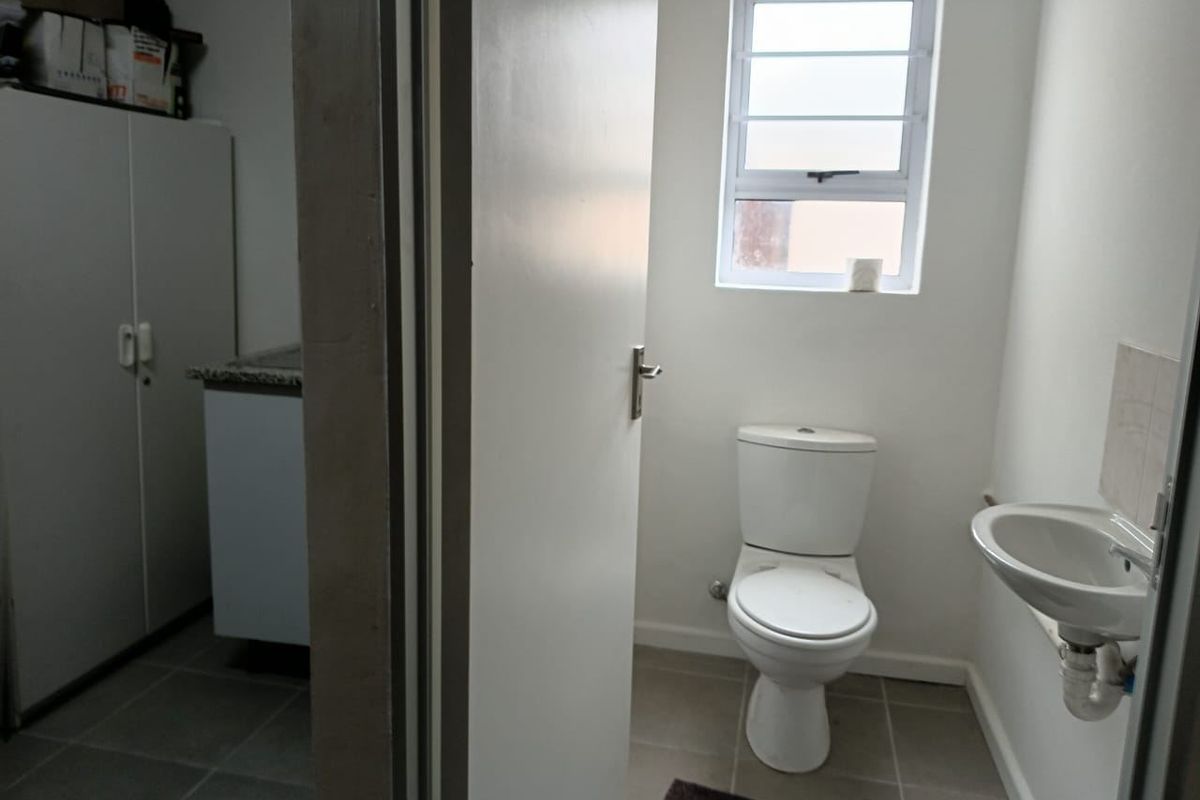 A functional bathroom featuring a standard toilet, wall-mounted basin, and tiled flooring, with a louvered window providing natural light.