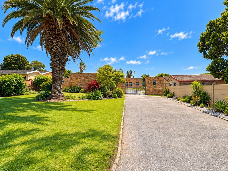A well-maintained front entrance featuring a paved driveway, secure metal gate, and established garden with mature palm trees.