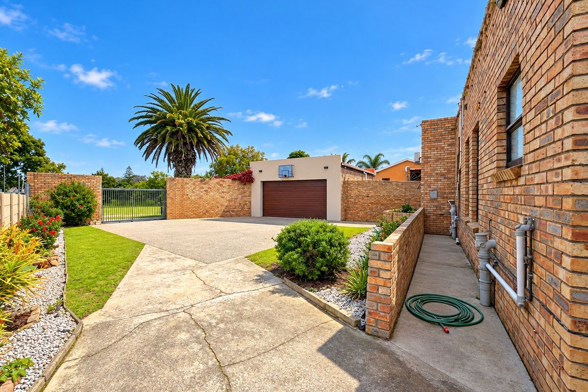 A well-maintained rear exterior featuring a brick facade, concrete driveway, single garage, and landscaped garden areas with a secure perimeter wall.