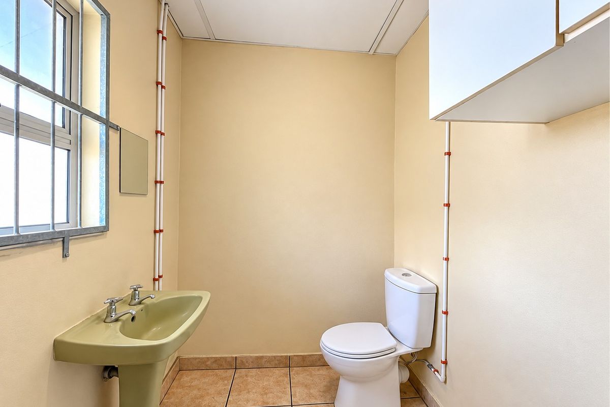 A functional guest bathroom featuring a pedestal basin, toilet, tiled flooring, and exposed surface-mounted plumbing pipes.