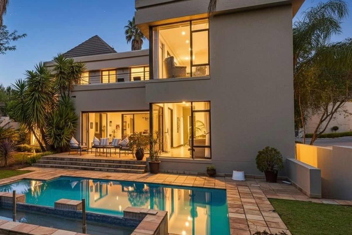 A beautifully illuminated rear exterior view featuring a modern two-story home, a sparkling swimming pool, and a spacious tiled patio area.