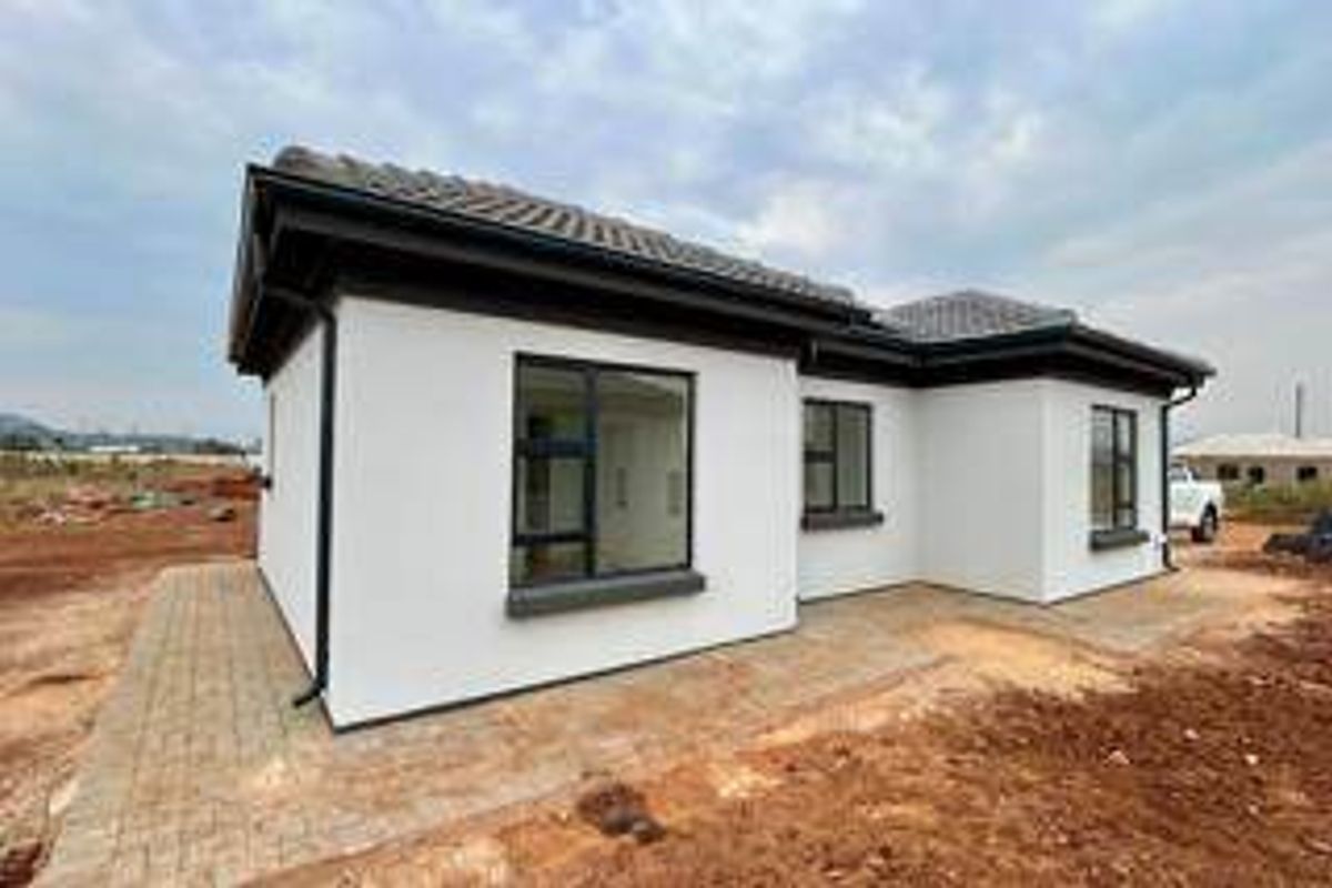 Rear view of a modern single-story house featuring white exterior walls, dark aluminium window frames, and a tiled roof with surrounding paved areas.