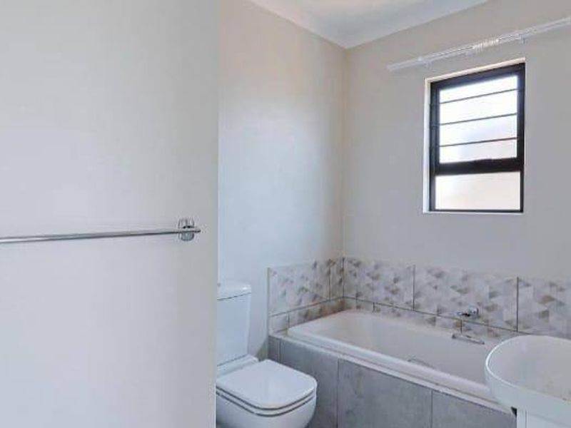 A clean and modern bathroom featuring a bathtub, toilet, basin, and a louver window, finished with neutral wall tiles.